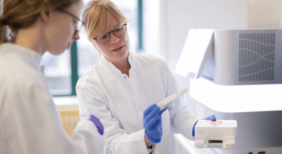 Two scientists in white lab coats and blue gloves work with equipment in a bright lab. They examine a sample, conveying focus and collaboration.